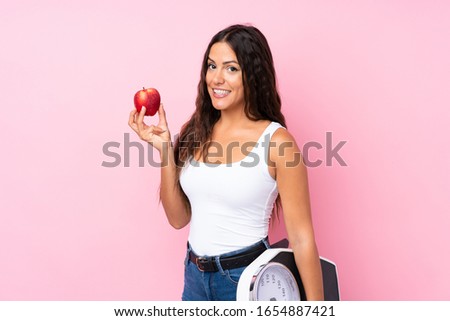 Young woman over isolated pink background with weighing machine and with an apple