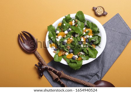 Blueberry, tangerine, cottage, feta, nuts and spinach salad served in a white bowl with handmade african wood cutlery. Denim fabric and yellow background.