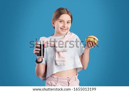 Portrait of positive teenage girl eating unhealthy hamburger and drinking cola against blue background