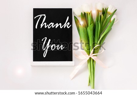 Thank You card message sign on black chalkboard with tulip flowers on white background flat lay. Blackboard greeting text top view. Natural floral decoration with green leaves. Thanksgiving Day banner