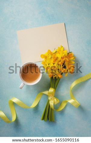 Morning cup of coffee, clean card and spring daffodil flowers on blue table. Beautiful breakfast for Woman day, Mother day. Top view.