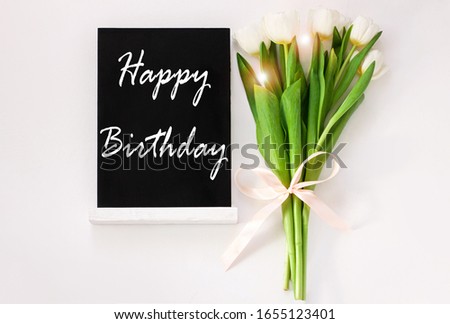 Happy Birthday card message sign on black chalkboard with tulip flowers on white background flat lay. Blackboard greeting text top view. Natural floral decoration green leaves. Celebrate party banner.