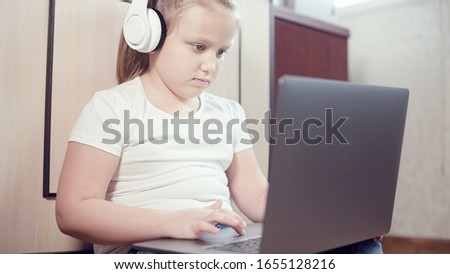 A smart little girl in white headphones with a laptop in her hands is pushing on the floor in her room. The young generation on the Internet and IT technology