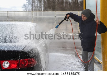 A picture of a man washing a car under high water pressure outdoors in the cold season.