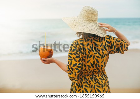 Picture of woman in beach dress look at ocean. Stand on beach or seashore with cocktail in coconut. Vacation mood and rest time. Calm peaceful woman and sunny beautiful weather