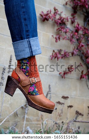 A close up of an elegant woman's leg wearing a beautiful wooden clog shoe with brown leather in front of a autumn leaves and limestone wall background Royalty-Free Stock Photo #1655151436