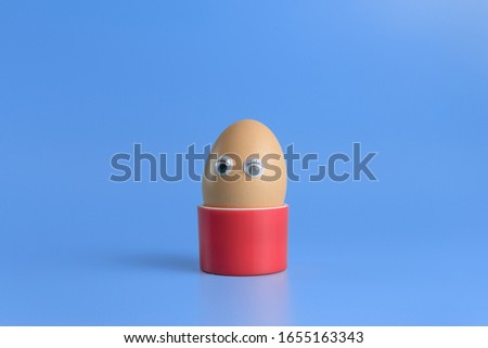 One brown egg with eyes in a round red box on a blue background. Minimal Happy Easter concept decoration. Copy space for text mockup. Close up photography.