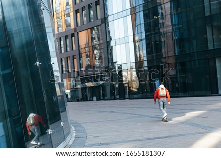 Trendy hipster in red jacket with green backpack riding on longboard among the modern houses. Urban and street photo. Healthy lifestyle, fashion look. Concept of leisure activity and youth.