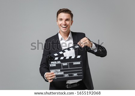 Cheerful young business man in classic suit shirt posing isolated on grey background. Achievement career wealth business concept. Mock up copy space. Holding classic black film making clapperboard