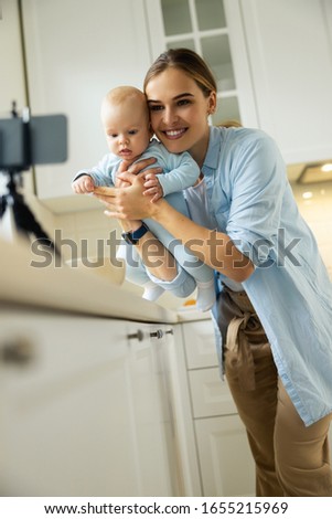 Smiling young mom holding little kid while using camera for blog in the kitchen stock photo