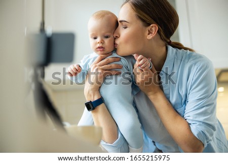 Smiling young mother holding little kid while using camera for blog in the kitchen stock photo