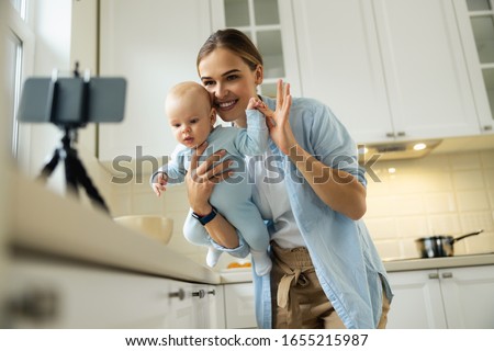 Smiling young woman holding little kid while using smartphone for blog in the kitchen stock photo