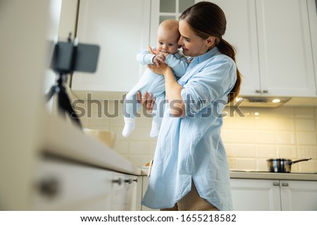 Smiling female holding cute kid while using smartphone for blog in the kitchen stock photo