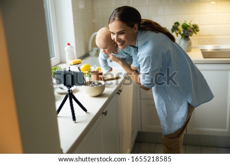 Smiling mom holding little kid while using smartphone for blog in the kitchen stock photo