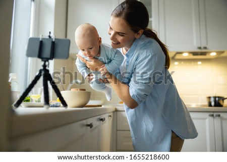 Happy lady holding child while using smartphone for blog in the kitchen stock photo