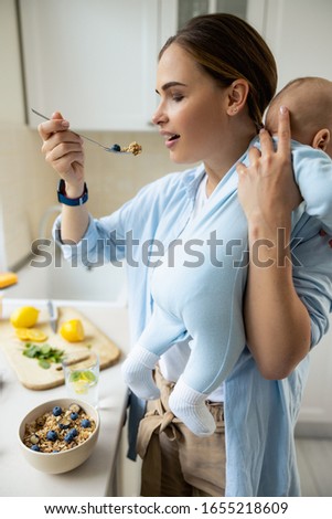 Pretty mother holding baby while eating her breakfast at home stock photo