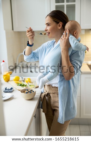 Pretty mom holding baby while eating her breakfast at home stock photo