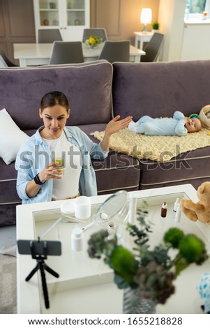 Top view of smiling pretty mother holding glass of water and using smartphone for blog stock photo
