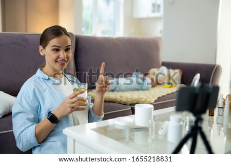 Smiling young mom holding glass of water and using mobile phone for blog stock photo