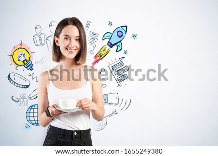Cheerful young woman in casual clothes drinking coffee near grey wall with colorful startup sketch drawn on it. Concept of business success. Mock up