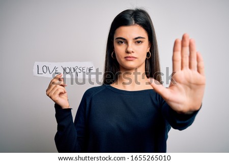 Young beautiful woman asking for take care of you holding paper with love yourself message with open hand doing stop sign with serious and confident expression, defense gesture