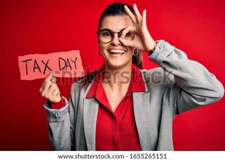 Young beautiful brunette businesswoman holding papaer with tax day message with happy face smiling doing ok sign with hand on eye looking through fingers