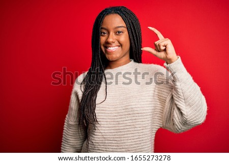 Young african american woman wearing casual winter sweater over red isolated background smiling and confident gesturing with hand doing small size sign with fingers looking and the camera. Measure 