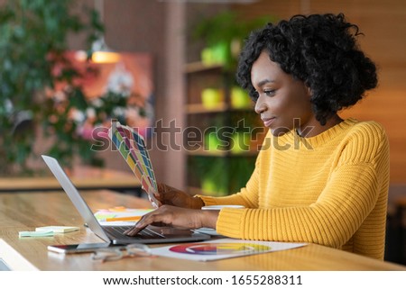 Creative afro girl web-designer working with color palette at cafe, using laptop, empty space