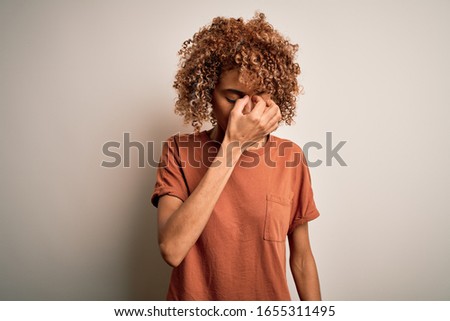 Beautiful african american woman with curly hair wearing casual t-shirt over white background tired rubbing nose and eyes feeling fatigue and headache. Stress and frustration concept.