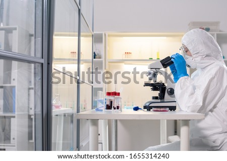 Side view of medical scientist in protective mask using microscope while studying dangerous coronavirus in laboratory Royalty-Free Stock Photo #1655314240