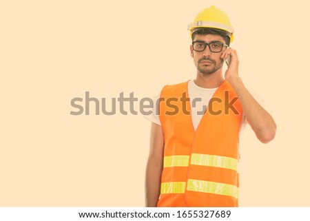 Studio shot of young Persian man construction worker talking on mobile phone