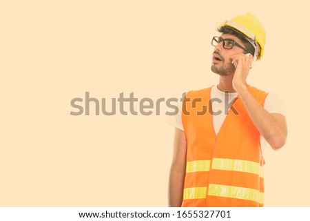 Young Persian man construction worker talking on mobile phone while thinking and looking up