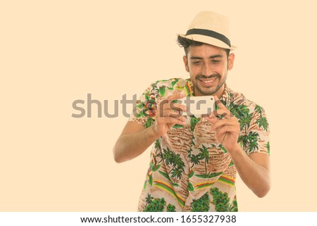 Studio shot of young happy Persian tourist man smiling while taking picture with mobile phone