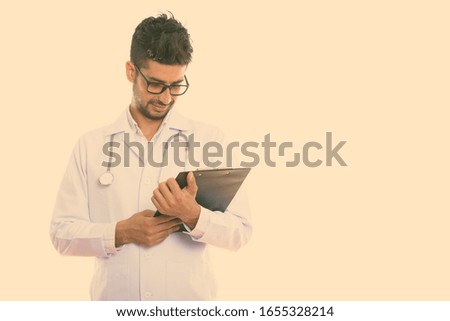 Studio shot of young Persian man doctor wearing eyeglasses while reading on clipboard