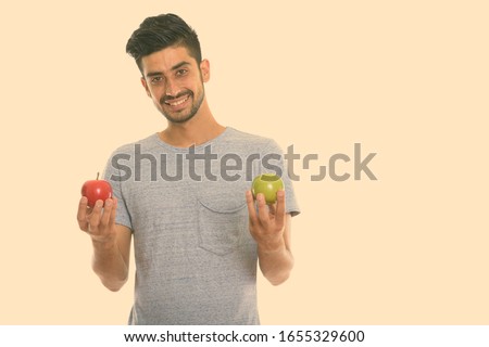 Studio shot of young happy Persian man smiling while holding red apple and green apple