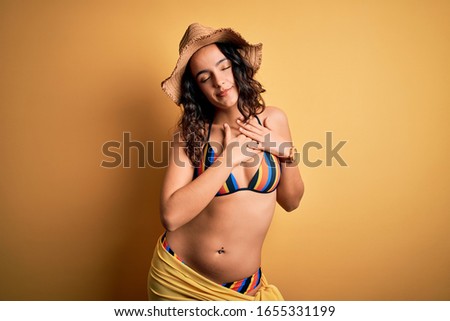 Young beautiful woman with curly hair on vacation wearing bikini and summer hat smiling with hands on chest with closed eyes and grateful gesture on face. Health concept.