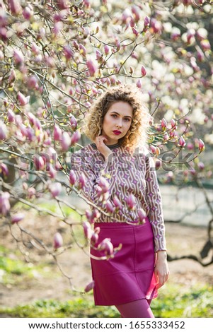 Portrait of blondy beautiful girl with curly long hair. Woman walks in the garden of blooming pink magnolia