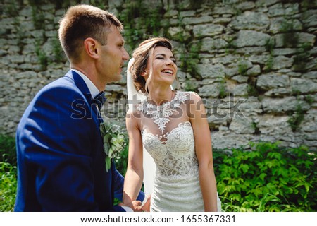 Beautiful bride holds the hand of the groom. Stylish newlyweds walking in the park outdoors. Loving couple on their wedding day. Portrait of smiling bride on stone wall background.