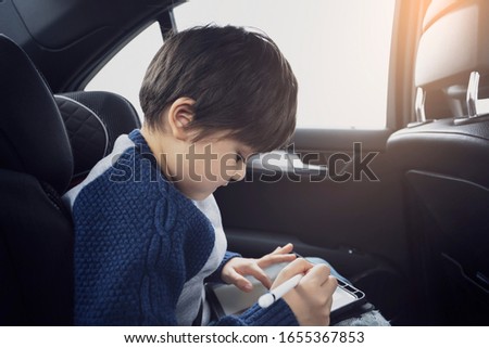 Happy young boy using a tablet computer while sitting in the back passenger seat of a car with a safety belt, Child boy drawing on smart pad,Portrait of toddler entertaining him self on a road trip