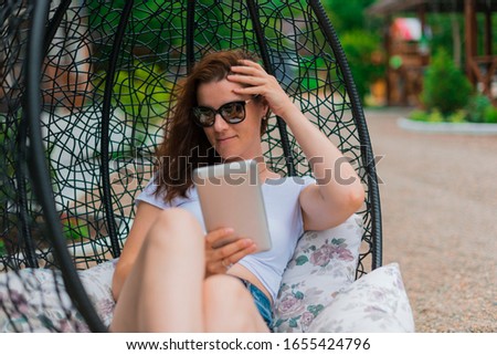 Young brunette girl in denim shorts white top black glasses. Resting in a chair on the outdoor in the hands of a tablet Communicates with friends via a modern gadget
