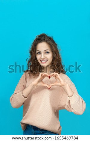 Portrait of a young beautiful woman wearing sweatshirt holding hands in the shape of a heart and looking at you coquettishly isolated over blue background