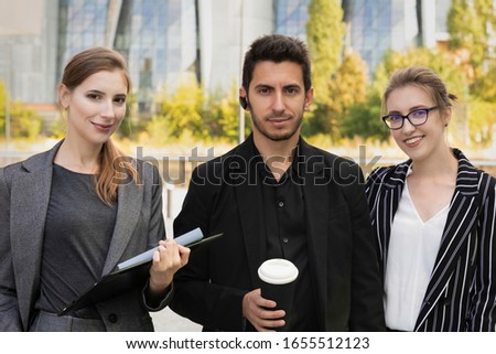 Three business partners stand against the background of an office building. They rejoice and smile, as they managed to successfully negotiate and conclude a deal.