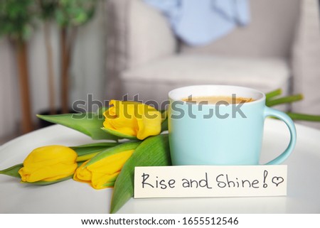 Aromatic morning coffee, beautiful flowers and card with RISE AND SHINE wish on white table indoors