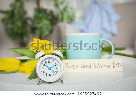 Alarm clock, aromatic morning coffee, beautiful flowers and card with RISE AND SHINE wish on white table indoors