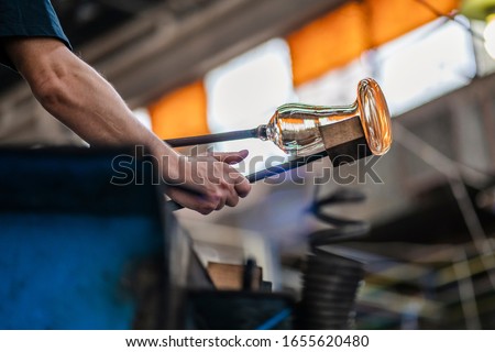 Caucasian glassblower artist is shaping the molten glass extracted from the furnace located in a glass factory in Romania where each piece is manually made by the artisans hands. Labour day. Royalty-Free Stock Photo #1655620480