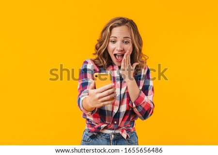 Image of young beautiful woman wearing plaid shirt rejoicing and holding cellphone isolated over yellow background