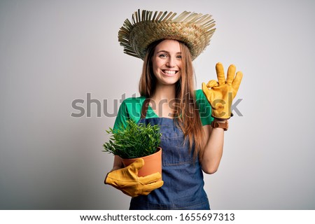 Young beautiful redhead farmer woman wearing apron and hat holding plant pot doing ok sign with fingers, excellent symbol