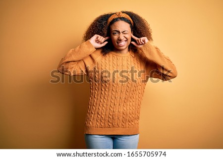 Young african american woman with afro hair wearing casual sweater over yellow background covering ears with fingers with annoyed expression for the noise of loud music. Deaf concept.