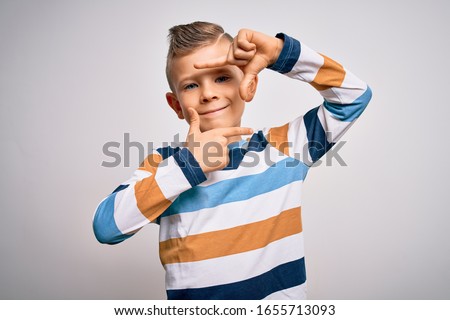 Young little caucasian kid with blue eyes standing wearing striped shirt over isolated background smiling making frame with hands and fingers with happy face. Creativity and photography concept.