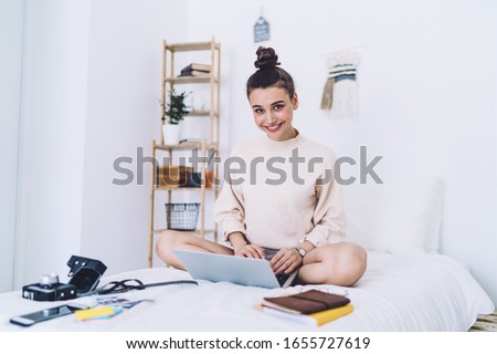 Casual young lady photojournalist with effulgent smile sitting legged cross on white sheets bed working with laptop and books in well appointed bedroom and looking at camera 